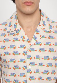 Kurzarmhemd aus leichtem Stoff, das ein wiederholendes Muster von blauen Kreaturen, grünen Blättern und roten Blumen auf einem beigen Hintergrund zeigt.