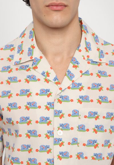 Chemise à manches courtes en tissu léger, arborant un motif répétitif de créatures bleues, de feuilles vertes et de fleurs rouges sur un fond beige.