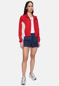 Giacca rossa corta con accentuazioni crema e blu, t-shirt bianca, pantaloni corti a righe blu navy e scarpe sportive argento.