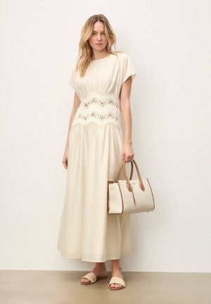 Vestido largo color crema con una cintura de encaje de crochet, combinado con un bolso beige y sandalias de punta abierta, de pie sobre un suelo neutro.