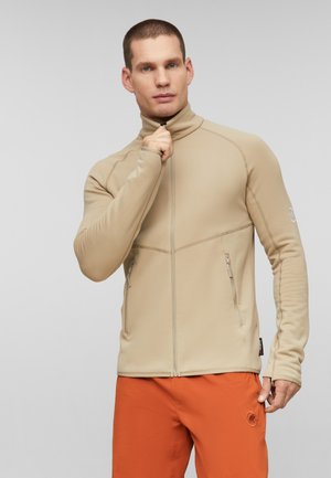 Uomo con giacca beige con zip e pantaloni arancioni, che tiene la zip della giacca vicino al collo su uno sfondo grigio uniforme.