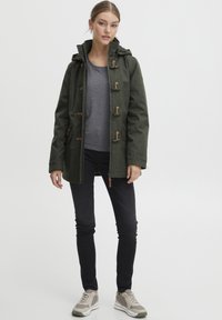 Forest green jacket with toggle closures, hood, and long sleeves, paired with a grey shirt and dark jeans, completed with beige sneakers.