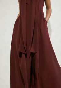 Ärmelloses weinrotes Kleid mit lockerem Schnitt, tiefen Taschen und langem Frontschal, der in der Mitte herunterhängt.