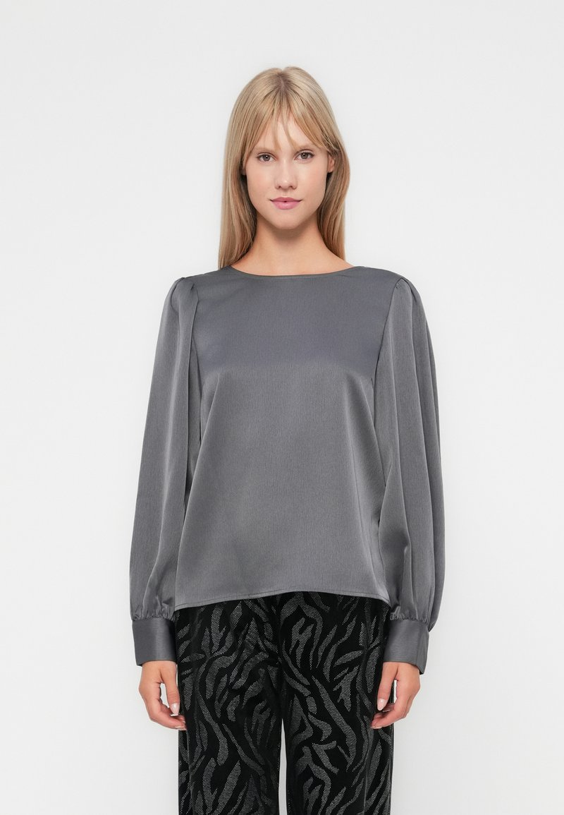 Maglietta grigia in satin a maniche lunghe con scollo tondo, caratterizzata da spalle abbondanti e polsini aderenti. Abbinata a pantaloni neri con motivo.