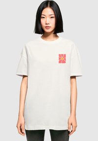 Hellbeige, oversized T-Shirt aus Baumwolle mit einem pinken und gelben Blumenmuster auf der linken Brustseite. Kurze Ärmel, runder Halsausschnitt.