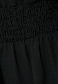 Schwarzer Stoff mit geraffter Smocking-Textur an der Taille, mit weichen, fließenden Drapierungen darunter. Das Material wirkt leicht und glatt.