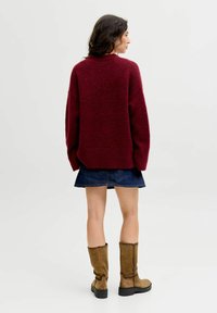 Burgunderfarbener übergroßer Pullover mit strukturiertem Strick, kombiniert mit einem Jeansrock und braunen kniehohen Stiefeln mit einer klobigen Sohle.