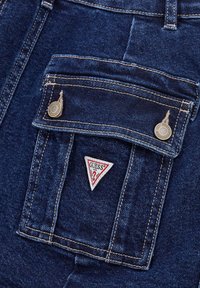 Des shorts en denim foncé présentent une poche avant avec une fermeture à bouton et un patch logo triangulaire "GUESS" en rouge et blanc. Couture contrastée visible.