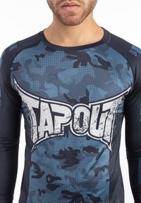 Hosszú ujjú sportpóló kék kamuflázs mintával, amelyen egy kiemelkedő fehér "TAPOUT" logó található, vastag élekkel és testhezálló fazonnal.