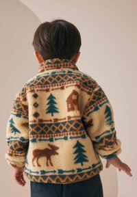 Enfant portant une veste en polaire beige ornée de motifs d'arbres verts, d'élans marron et de maisons, se tenant dos à l'appareil photo devant un fond uni.