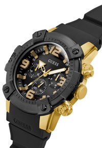 Orologio nero e oro con cassa rotonda, cinturino in gomma, numeri grandi, tre sotto-quadranti per cronografo e accenti testurizzati sulla lunetta.