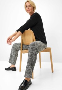 Zwarte trui, gestructureerde grijs-witte gebreide broek, zwarte instappers, zittend op een houten stoel tegen een witte achtergrond.