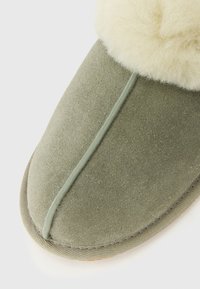 Groene suede pantoffel met crèmekleurige fleece voering en rubberen zool, met close-up van het teengebied.