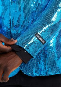 Sequin mėlyna striukė išsiskiria blizgiąja tekstūra ir kontrastinėmis juodomis rankovėmis. Ant rankovės matoma etiketė su užrašu "SUTIMEISTER."