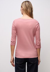 Rosa, langärmliges Oberteil mit einem Boots-Ausschnitt, das eine glatte Textur aufweist. Pullover-Stil, kombiniert mit dunklen Denim-Hosen.