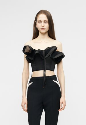 Ung kvinde med langt, glat hår iført sort, rynket crop top med bare skuldre og sorte, højtaljede bukser med hvide detaljer.