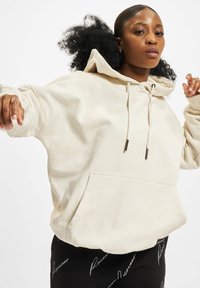 Beige, oversized kapucnis pulóver zsinórokkal és elülső zsebbel, finom fehér mintákkal. Fekete nadrággal párosítva, amelyen fehér szöveges részletek találhatók.