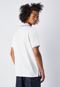 Weißes Polohemd mit kurzen Ärmeln, schwarzer Einfassung am Kragen und an den Bündchen sowie seitlichen Schlitzen. Sanfte Textur, klassisches Design und lässige Passform.