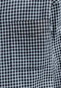 Ύφασμα με καρώ μαύρο και ανοιχτό μπλε χρώμα με μικρό τσεπάκι στο στήθος· διαθέτει καθαρές γραμμές και λεία υφή.