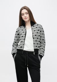 Black and white zip-up jacket with repeated logo pattern, ribbed cuffs, and collar. Paired with black pants featuring side stripes.