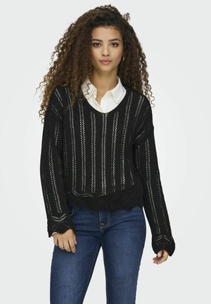 Maglione nero in maglia con righe verticali, orlo scallop e vestibilità ampia. Indossato sopra una camicia bianca con colletto, dotata di maniche lunghe.