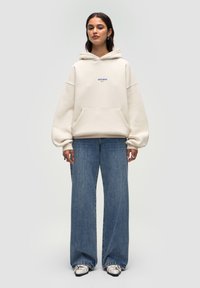 Cremfarbener Hoodie mit einer vorderen Kängurutasche und Logo, kombiniert mit hellblauen weit geschnittenen Jeans und schwarz-weißen Schuhen. Glatte Textur.