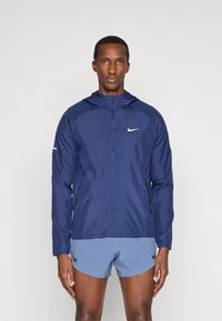 Navy blue Nike windbreaker with a zipper front, adjustable hood, and lightweight fabric. Features a white logo on chest and contrasting accents.