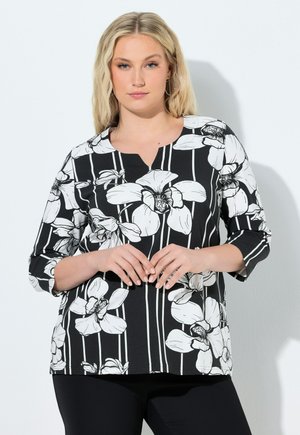 Lyshåret kvinde iført sort-hvid blomsterprintet langærmet top og sorte bukser, stående foran en hvid baggrund.