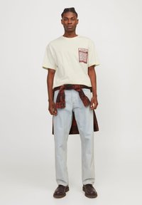 T-shirt beige clair avec une poche à motif rouge, associé à un jean bleu clair à jambe droite et une chemise à carreaux sombre nouée à la taille. Chaussures marron.