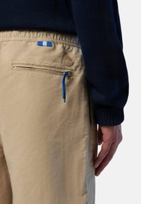 I pantaloni beige presentano una tasca con zip dotata di un tirante blu e un accento con logo circolare. La cintura ha un design elastico per il comfort.