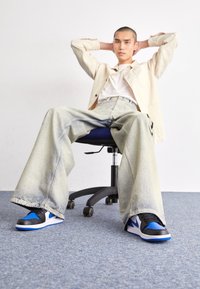 Černé a modré tenisky s plochou podrážkou, spárované se širokými, světlými džíny. Osoba sedí na otočné židli na modrém koberci.