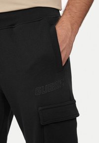 Pantaloni della tuta neri con tasca frontale, caratterizzati da un logo testurizzato "GUESS". Il tessuto è liscio con una leggera lucentezza, che enfatizza una vestibilità moderna.
