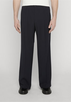 Mann trägt locker sitzende dunkelblaue Hose und schwarze Anzugschuhe, steht vor einem schlichten hellen Hintergrund.