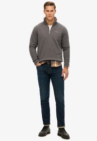 Suéter gris con cremallera y cuello de cuadros, superpuesto a una camisa blanca. Combinado con jeans azul oscuro y zapatos negros. Look limpio y casual.