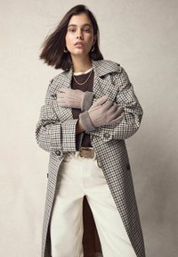 Rute trenchcoat i beige og blå, lagdelt over en brun sweater, parret med lyse jeans og grå handsker med håndledsdetaljer.