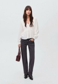 Kvinna med långt mörkt hår som bär en vit blus, mörka jeans, svarta stövlar och håller en mörkröd handväska mot en enfärgad bakgrund.