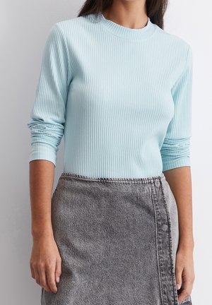 Vrouw die een lichtblauw geribd shirt met lange mouwen draagt, ingestopt in een grijze denim rok met hoge taille.