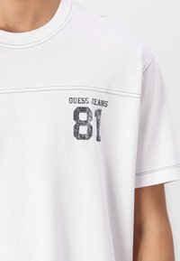 Hvit bomull t-skjorte med rund hals, korte ermer og en trykket svart "GUESS JEANS 81" logo på forsiden med kontrasterende søm.