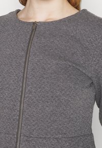 Grå fleeceplagg med rund halsringning och en central dragkedja. Texturen är mjuk, med släta sömmar och en figursydd siluett.