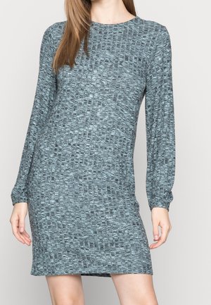 Mujer con un vestido gris ajustado de manga larga y textura, con escote redondo, de pie sobre un fondo liso y claro.