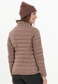 Femme souriante, portant une doudoune mauve, un bonnet en tricot marron et un pantalon noir, de profil sur un fond blanc uni.