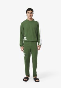 Felpa verde e pantaloni abbinati, con accenti bianchi e un logo a forma di coccodrillo. Il materiale appare morbido. Indossato con sandali neri.
