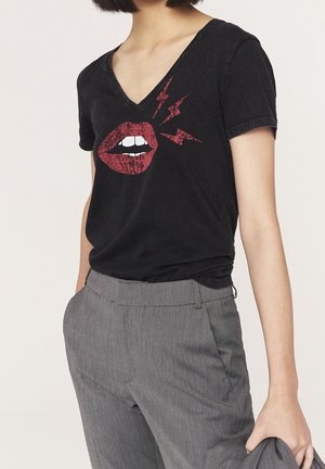 Personne portant un t-shirt noir à col en V avec une bouche rouge graphique et trois éclairs, associé à un pantalon gris texturé, tenant un tissu dans la main.