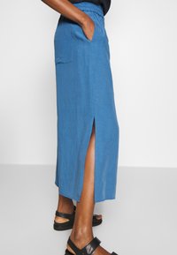 Lichtblauwe enkellange rok van zachte stof, met een elastische tailleband en een zijsplit, gecombineerd met zwarte sandalen.