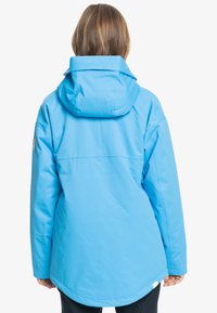 Blå vattentät jacka med huva, med en slät yta, förlängd bak och minimalistisk design, perfekt för utomhusaktiviteter.