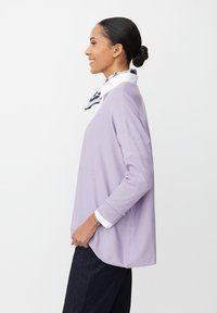Maglione viola a maglia con maniche lunghe e vestibilità ampia, indossato sopra una camicia bianca con colletto a righe navy. Texture liscia, design casual.