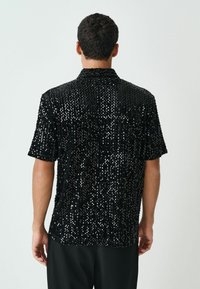 Schwarzes Kurzarmhemd, das mit Pailletten bedeckt ist, mit Kragen und legerem Schnitt. Glatte Textur mit glänzendem Finish.