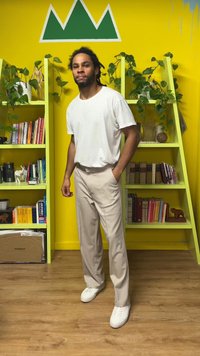 Model v beli majici in bež hlačah, stoji na lesenem tleh, za njim so zeleni regali in rumeni zid. Na nogah so vidni beli supergi.