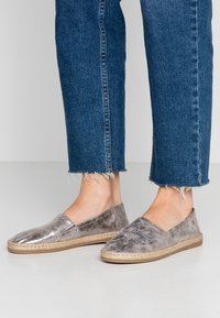 Pies con zapatos plateados metálicos tipo mocasín con suelas trenzadas y jeans azules con dobladillo deshilachado, sobre un fondo liso.