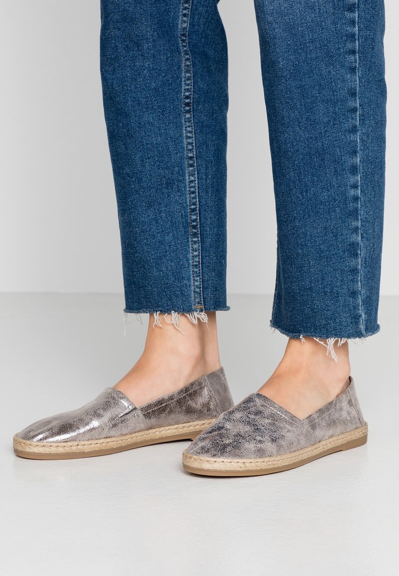 Pies con zapatos plateados metálicos tipo mocasín con suelas trenzadas y jeans azules con dobladillo deshilachado, sobre un fondo liso.